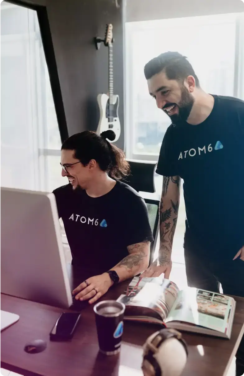Dois homens sorridentes, ambos com camisetas pretas da 'ATOM6', interagindo em frente a um monitor de computador. Um homem com cabelo preso em um coque está sentado, enquanto o outro está em pé, apontando para a tela.