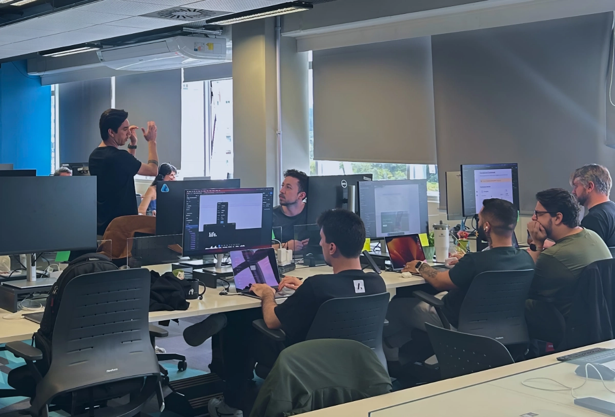 Um grupo de desenvolvedores em um escritório aberto, alguns sentados em suas mesas com computadores, enquanto um homem de camiseta preta gesticula e fala em pé, conduzindo uma discussão ou apresentação.