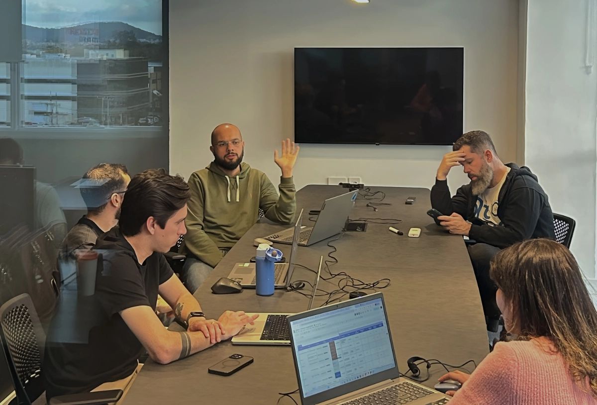 Um grupo de cinco pessoas em uma sala de reunião com uma mesa grande. Um homem com barba e óculos, vestindo um moletom verde, gesticula com a mão levantada, enquanto os outros membros da equipe olham para laptops ou celulares.